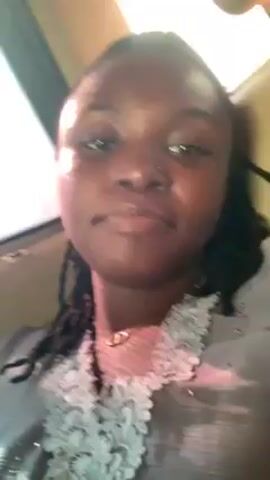 The video shows a woman with dark skin, dark eyes, and braids. She is wearing a gray top with lace detailing around the neckline and a necklace with a peace sign charm. She has a nose ring in her left nostril. She is sitting in a car, as evidenced by the window and interior visible behind her. The lighting is bright, causing some glare on her face. She looks directly at the camera with a neutral expression.
