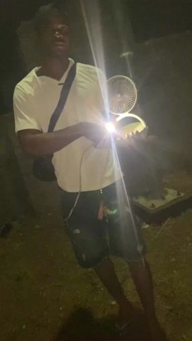 A person is outside at night. They are holding a small flashlight in one hand and a portable fan in the other, with the fan lit up from behind. The person is wearing a white t-shirt, dark shorts, and a dark baseball cap, and they have a bag slung across their shoulder. The lighting is poor except for the lights from the flashlight and the fan, so the surroundings are mostly dark and indistinct. The person moves slightly while holding the items.