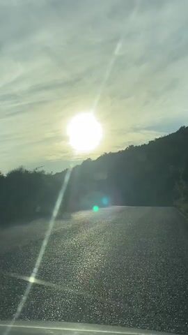 The video shows a view from a car driving on a paved road towards a bright sun peeking over a forested hill. The sun is a large, glowing orb in the sky, casting a bright light and creating lens flares on the camera. The sky is partly cloudy, with thin streaks of white clouds against a light blue backdrop. The road surface is dark and appears wet or reflective, with small pebbles visible. Green foliage and trees line the sides of the road and cover the hill. The overall impression is a scenic drive during sunrise or sunset.