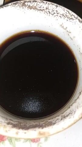 A close-up, overhead shot shows a white ceramic mug filled with a dark liquid, likely coffee. The mug has a textured rim with brown speckling. The liquid's surface is smooth and reflects light. Tiny particles are visible suspended in the liquid and scattered on the mug's inner surface. The background is blurred, suggesting a tabletop.