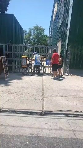The video shows an outdoor scene on a sunny day. An ice cream vendor on a bicycle is pulling his cart through a metal gate while two people stand nearby. A man in a white hat and shorts walks through the gate after the ice cream vendor. To the left, there is a food stand with a chalkboard menu next to a green storage container. A person wearing an orange shirt sits at the food stand. Trees can be seen in the background. A large building with green panels is on the right. There are power lines along the bottom of the frame.