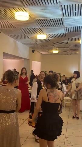 Several women dance in a room decorated for a wedding or similar formal event. Most of the women wear dresses. One woman wears a folk costume with a patterned vest, long skirt, and green headband. The room has white walls, a white and brown tile floor, and a white ceiling with round lighting fixtures. Tables with white tablecloths are arranged in the background, occupied by seated guests. Confetti litters the floor. The women laugh and raise their hands in unison, dancing to music that plays in the background.