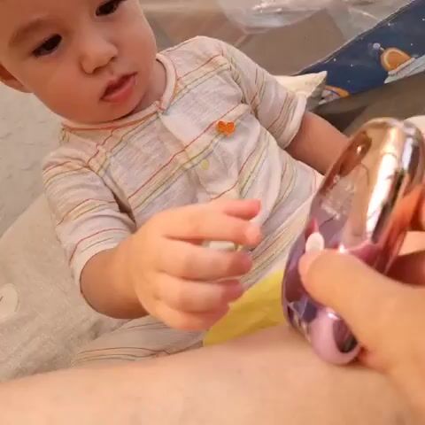 The media file shows a close-up shot of a baby and a person's hand holding a pink and gold hair clipper. The baby, wearing a striped onesie with orange accents, is looking at the clipper with curiosity. The person's hand is interacting with the clipper, seemingly demonstrating its use or allowing the baby to explore it.

The setting appears to be indoors, possibly a home, with soft lighting suggesting daytime. The background is slightly blurred, but some elements like a textured surface and a colorful patterned fabric are visible, hinting at a domestic environment. The focus is on the interaction between the baby and the hair clipper.

The baby's expression is one of interest and engagement, while the person's hand is gently guiding the object. There are no explicit indications of the time of day or weather. The location context provided places the scene in Dunnae-myeon, South Korea.

Visible text on the hair clipper includes branding that appears to say "Kemei" and various certification marks like "CE" and "FCC." There is also a QR code and some smaller Korean text. The overall mood is calm and inquisitive, centered around a child's exploration of a common household item.