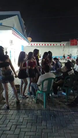 A group of people are gathered outdoors at night. Several young women in short dresses are standing and interacting with each other and the men present. Some men are seated at tables in the background. The area appears to be paved and decorated with red and white bunting.
