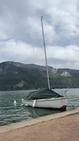 A stationary sailboat, covered by a green tarp, is docked along a concrete edge by a lake. The hull is white with a red stripe at the bottom. A tall, silver mast extends upward. White buoys float in the water nearby. A backdrop of mountains is visible under a cloudy sky. The water ripples gently.