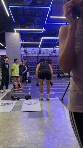 The video shows a group of people in a gym. In the foreground, a woman with long dark hair is holding a phone to her ear and looking to the left. She is wearing a sleeveless top and black leggings. To her right, a man in a black tank top and shorts is performing a dumbbell exercise. He is facing away from the camera, bending at the waist, with a dumbbell in each hand. He is on a mat on the floor. Behind him, there are other people standing and watching. Two men and one woman are standing to the left of the man exercising. One man is wearing a black t-shirt and pants, another man is wearing a green t-shirt and shorts, and a woman is wearing a dark tank top and shorts. In the background, there are gym equipment and blue neon lights on the ceiling. The gym appears to be a modern facility.