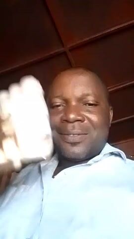 A man is holding a group of small, white, plastic containers wrapped in a plastic bag above his head. He is smiling and looking directly at the camera. He is wearing a light blue button-down shirt and has a wedding band on his left ring finger. The background is a brown, wooden structure. A partial box with red and blue lettering is visible behind the man.