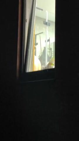 A person is visible through a window, appearing to be in a shower. The window is partially open, revealing a bathroom with a shower stall. The lighting is dim, and the person's face is not clearly visible. The person's back is towards the viewer, and they appear to be wearing a light-colored garment. The environment outside the window is dark, suggesting it is nighttime.
