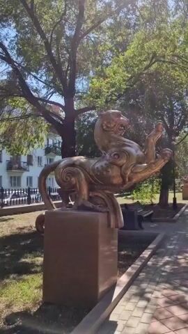 A golden statue of a mythological creature, resembling a lion or dragon, is positioned on a stone pedestal in an outdoor setting. The statue is ornate, with stylized swirls and sharp claws. In the background, a light-colored, multi-story building with balconies and windows is visible. Trees with green foliage provide a canopy above, and a paved walkway runs along the foreground. The sky is clear and blue, suggesting a daytime scene. There are no people or animals directly interacting with the statue, though a park bench can be seen in the distance.