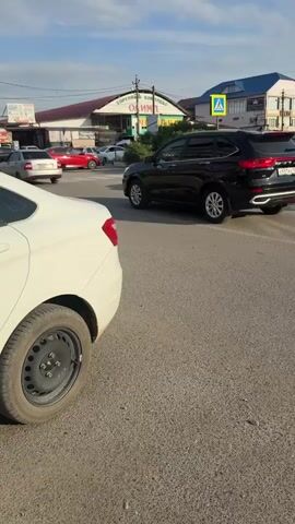 The video shows a street scene in Kizlyar, Russia, during the daytime. The sun appears to be setting, casting a warm light on the buildings and cars. The weather seems clear with a bright blue sky dotted with some clouds.

The main subjects are several cars on a paved road. In the foreground, a white car is partially visible from the rear passenger side, showing its wheel and part of the bumper. Further down the road, a black SUV is the most prominent vehicle, facing away from the camera and appearing to drive off. To the right of the SUV, two white sedans are parked or moving slowly. A black hatchback is also visible in the middle ground, driving in the same direction as the SUV. In the background, more cars are seen, including a red sports car.

Behind the cars, there are several buildings. A prominent building has a sign with Cyrillic text that appears to read "ТОРГОВЫЙ КОМПЛЕКС ОЛИМП" (Trading Complex Olimp). Another building to the right has a blue roof and some architectural details. A yellow pedestrian crossing sign is also visible, indicating a street intersection.

The activity captured is the movement of vehicles on a street, likely in a commercial area given the "Trading Complex" sign. The overall impression is a snapshot of everyday life and traffic in a Russian town during the late afternoon or early evening. There are no people clearly visible. The mood is neutral, focusing on the urban environment and vehicular traffic.