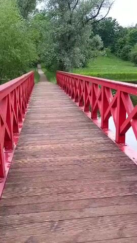 This video shows a POV shot of a person walking across a wooden bridge with bright red railings. The bridge spans over a body of water. Lush green trees and grass line the sides of the bridge and the surrounding landscape. A path is visible leading away from the bridge into the greenery. The weather appears to be overcast.