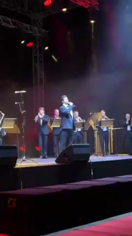A man in a dark suit sings onstage while a small band accompanies him. The band, also in dark suits, consists of wind instruments like trumpets, clarinets, and a saxophone. They're all standing at music stands, playing in front of microphones. The stage is lit with red and yellow lights. There's a black speaker system at the front of the stage. The background features a metal truss structure with more lights. In the distance, there are blurred hints of an audience.