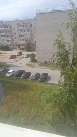 The video shows a view of an apartment complex courtyard on a sunny day. Several cars are parked in a lot and along a curved road. A satellite dish is visible in the foreground. The buildings are made of concrete and brick, with many windows. Green grass and trees are scattered throughout the area. No people are visible.