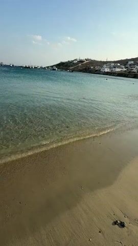 The media file displays a tranquil beach scene at Agios Ioannis Diakoftis, Greece. The foreground features a sandy beach with gentle waves lapping at the shore. The clear turquoise water allows a view of the sandy bottom beneath. In the mid-ground, the sea is dotted with numerous sailboats and yachts anchored offshore. Beyond the water, a coastal landscape unfolds, characterized by hills dotted with white buildings. The sky above is a clear, bright blue with a few wispy clouds, suggesting fair weather and a daytime setting, likely late afternoon given the warm light. The overall atmosphere is one of peace and relaxation. There are a few people visible in the water, enjoying the calm sea. The scene evokes a sense of summer vacation and a serene coastal environment. No text is visible in the media file.