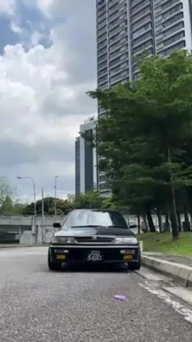 The camera focuses on a black Honda car, model and year not possible to determine, parked on the side of a grey asphalt road. The car is facing forward, and its license plate reads “JDD 3480”. A small, violet-colored object is on the road slightly in front of the car. Behind the car, there is a modern high-rise building, grey in color. To the right of the car are green trees, and on the left are more trees and a concrete overpass. The sky is blue with white clouds. The video appears to be taken during the day in an urban environment.
