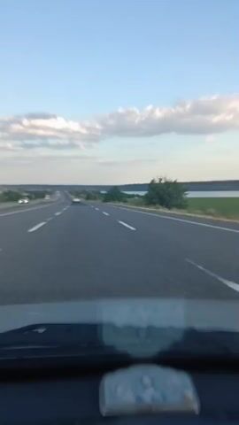 A first-person perspective video shows a view from inside a car driving on a highway. The road ahead is clear, with white lines marking the lanes. In the distance, several other vehicles are visible, as well as trees and a body of water on the right side of the road. The sky is blue with scattered white clouds. On the dashboard, there is a small religious icon.