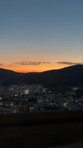 This is a vertical, handheld video of a sunset over a city nestled between hills. The sky transitions from a deep blue at the top to shades of orange and pink near the horizon. The city below is illuminated by numerous lights, suggesting it is dusk or evening. The foreground is dark, obscuring details. The camera appears to be moving, possibly from a vehicle, as indicated by the slight motion blur and the way the landscape shifts. The scene is peaceful and scenic, capturing the beauty of a city at twilight.