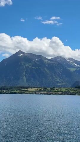 This media file captures a scenic view of Lake Thun in Switzerland on a clear, sunny day with fluffy white clouds scattered across a vibrant blue sky. The foreground is dominated by the rippling blue water of the lake, with a visible white wake trailing from the perspective of a moving boat in the lower left corner.

In the middle ground, the opposite shore of the lake is lined with lush green trees, forming a dense forest that stretches across the landscape. Beyond the treeline, rolling hills dotted with small villages and farmhouses suggest a rural setting.

The background is defined by a majestic mountain range, with the peaks partially covered in snow, indicating a significant elevation. The mountains are a deep green and brown, showing the contours of the terrain. Sailboats can be seen gracefully navigating the lake, adding a sense of calm activity to the serene environment.

The time of day appears to be daytime, likely late morning or early afternoon, given the bright sunlight illuminating the scene. The weather is pleasant and fair, conducive for outdoor activities. The overall mood is tranquil and picturesque, highlighting the natural beauty of the Swiss landscape. There is no visible text in the media file.