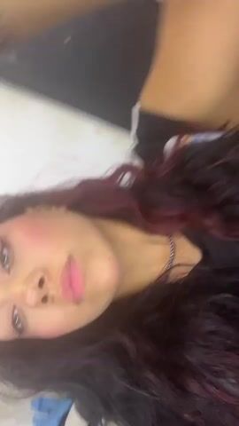 A person with dark wavy hair and some red streaks, wearing a black shirt with white trim, poses for a selfie. Their face is turned towards the camera, and their right arm is raised behind their head. Their makeup includes dramatic eyeliner and pink blush. The background appears to be an indoor setting with tiled flooring and a wall. The person sticks their tongue out briefly at one point.