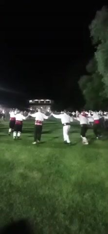 A group of people are dancing in a circle on a grassy field at night. They are holding hands and moving in a synchronized manner. Most of the dancers are wearing white shirts and red sashes. Some are also wearing dark pants or skirts. There are trees in the background, and some lights are visible in the distance. The overall atmosphere appears festive.