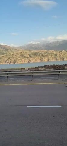 A white SUV with a black roof rack is seen driving on a road next to a body of water and mountains. The SUV appears to be a van or a similar passenger vehicle. The camera perspective is from another vehicle, possibly a truck, as a blue truck bed is visible in the foreground. The road has a yellow line and white lane markers. The sky is clear with some clouds. The mountains in the background are dry and brown, with some greenery. The water is calm and blue. The scene appears to be a scenic landscape, likely a highway or a scenic route.