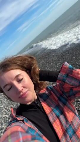 A woman with auburn hair stands on a rocky beach, holding her head with her hand and looking directly into the camera. She is wearing a black shirt underneath a bright, colorful plaid jacket. Behind her, waves crash on the shore, and a clear blue sky is visible with scattered clouds. Mountains appear in the far background.