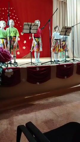 On a stage decorated with red fabric, five young girls stand in front of microphone stands, sheet music on stands in front of them. The girls are dressed in colorful skirts. Two of the girls wear green t-shirts, one a red shirt, and two white t-shirts. They are singing together. The stage is decorated with fake flowers. The recording appears to have been taken from the audience.