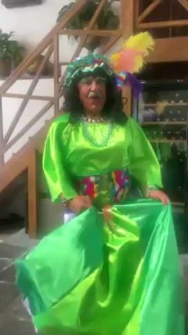 The video shows a person in a green, satin, oversized dress, a colorful belt, and a flamboyant headdress with feathers, likely a costume, dancing in an open-air courtyard setting with a stone floor.  The person spins and moves towards a wooden staircase and passes by a large red floral arrangement.  In the background, there is a display case, wooden beams, and potted plants. As the camera pans, a few other people can be seen including a server carrying something on her head and a man in a red mariachi suit. The video appears to be taken in a restaurant or event space.