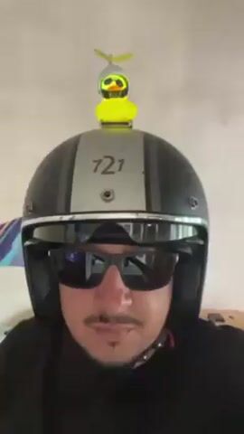 A man wearing a black t-shirt, sunglasses, and a matte black and silver motorcycle helmet with a short visor is seen from the chest up in an eye-level shot. The helmet has a small, lit-up yellow rubber duck with a propeller attached to the top. The duck alternates between emitting a bright red and a soft yellow glow. The background appears to be an interior space with light-colored walls and a partially visible colorful painting or textile hanging.