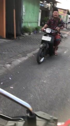 A man is riding a motorcycle on a paved street. He is wearing a red and black patterned shirt and a blue and white sarong. He is also wearing a black skullcap. The motorcycle is black. The street is lined with buildings and vegetation. The man is smiling and looking at the camera. The video is shaky and out of focus.