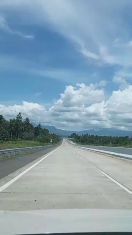 The video shows a view from inside a vehicle traveling on a highway. The highway is paved with concrete and has white lines marking the lanes. On the left side of the highway, there is a metal guardrail and then a roadside with trees and vegetation. On the right side, there is a concrete barrier. The sky is blue with fluffy white clouds, and in the distance, there are mountains covered in green vegetation. The scene appears to be a scenic drive through a rural or mountainous area.