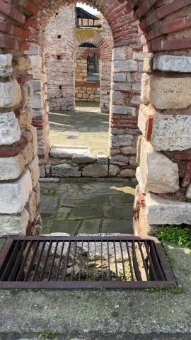 The video shows a static, low-angle view of a stone and brick structure, possibly ruins. The structure features a series of arched openings creating a corridor or passageway. The walls are made of weathered stone and brick, with some bricks showing a reddish hue. The floor is paved with stone slabs, some showing signs of moss or weathering.

In the foreground, there's a metal grate set into the floor. Beyond the first arch, the view continues through more arched openings, creating a layered perspective. Sunlight shines through the openings, casting shadows on the floor.

In the distance, through the arches, a building with a traditional roof design is visible. The scene is peaceful and quiet, focusing on the architectural details and the play of light and shadow within the structure.