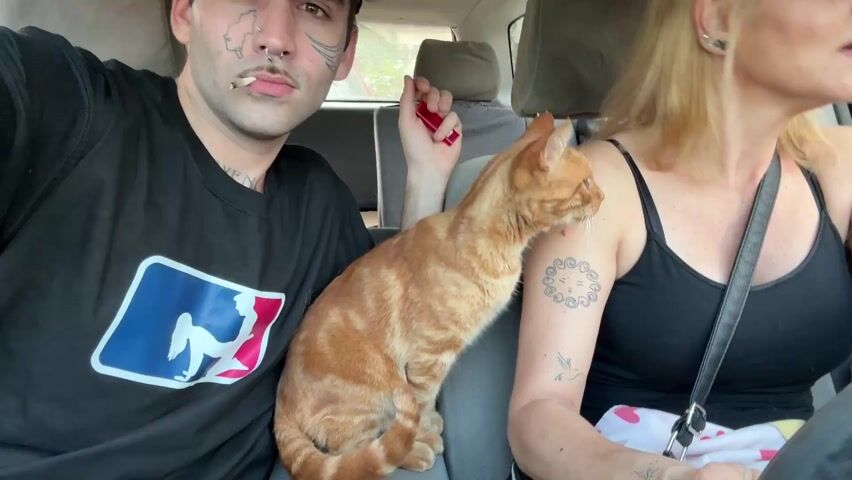 A video shows the inside of a car, where two people and a cat are seated. A young man is sitting in the passenger seat. He is wearing a black t-shirt with a baseball logo on it, and a red baseball cap. He has tattoos on his face and neck, and a cigarette is in his mouth. He is holding a red lighter in his hand. Beside him is a fair-skinned woman in a black tank top. She is the driver, and appears to be addressing the man in the passenger seat. Between the two people is an orange tabby cat. The car's interior is gray. The man looks at the camera in the beginning and continues looking as the woman talks.