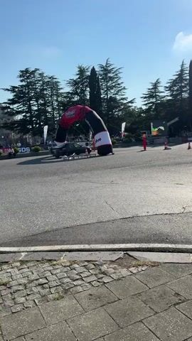 A dark-colored SUV is seen driving through an inflatable archway with red and black checkered patterns on a paved road. The archway has the word "DRIVE" printed on it. There are orange traffic cones on the road, and a large screen displaying colorful graphics is visible in the background. Several people are standing around the area, some appearing to be spectators. Tall cypress trees line the background, and the sky is clear and blue. The ground in the foreground is paved with cobblestones. A banner with writing is visible behind a line of cones.