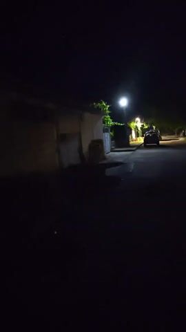 This is a nighttime video of an empty street with a street light on. There is a car parked on the side of the road and some trees in the background. The street is paved and there is a sidewalk on one side. The video is shaky and the lighting is poor.