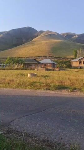 A scenic view of a rural landscape at sunset or sunrise. Rolling hills bathed in golden light dominate the background. In the mid-ground, a few houses are visible, nestled amongst dry grass and vegetation. A paved road cuts across the foreground, with a patch of green grass and some dry weeds on the side. The sky is clear and blue, with the sun low on the horizon casting long shadows.