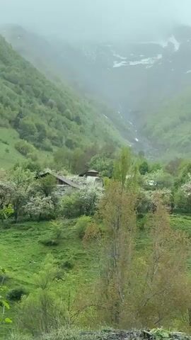 A video shows a lush, green mountainous landscape. The valley is filled with trees and a small village is nestled among them. A winding path leads down to a stream that flows through the valley. The mountains are partially covered in mist, and there are patches of snow visible at the higher elevations.