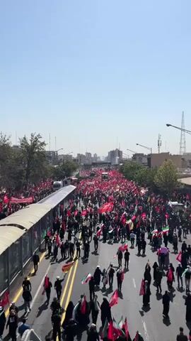 A high-angle, vertical video shows a large crowd of people walking down a wide street. The majority of the people are wearing dark clothing. Many are holding flags, predominantly red, with some green, white, and black flags also visible. The street is lined with trees on the left and buildings on the right. Several large trucks are visible interspersed within the crowd. The sky is clear and blue. The overall impression is of a procession or demonstration.