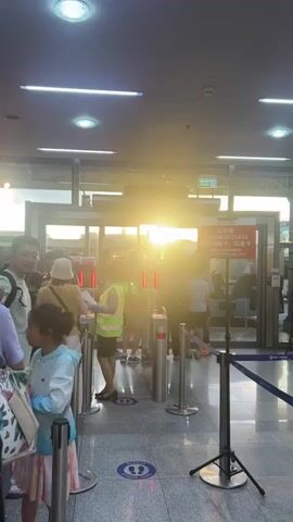 A group of people are queuing at a set of automatic gates. The sun is shining brightly through the gates, creating a lens flare effect. A woman wearing a yellow vest appears to be scanning something for the people in line. There are signs with Chinese writing above the gates. The flooring is tiled, and there are stanchions with ropes guiding the queue.