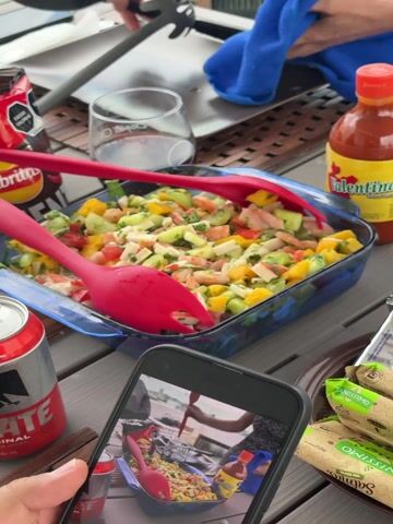 The video shows a close-up of a blue glass dish filled with a shrimp and mango salad, being served with red salad tongs. The salad contains chopped mango, shrimp, cucumber, and imitation crab meat. The dish sits on a gray wooden table, surrounded by items including a bag of chips, a bottle of Valentina hot sauce, a can of Tecate beer, a glass, and packages of crackers. Someone is using a smartphone to photograph or record the scene. A blue cloth is visible in the background, held by a person whose hand is in the frame.
