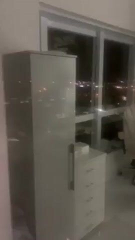 The video shows a room at night. There is a large window with a view of city lights. Next to the window are several pieces of furniture, including a tall cabinet, a chest of drawers, and an office chair. At the end of the video, a medical cart with lights and tubing comes into view.