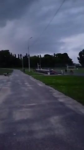A person is walking on a paved path next to a grassy field. In the distance, there is a soccer field with a soccer goal, several people, and a few bicycles. The sky is overcast and gray.