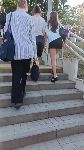 The video shows a group of people walking up a set of outdoor stairs. The subjects include at least four individuals, seen from the waist down, ascending the steps. One person in the foreground, wearing a striped shirt and black pants, carries a black bag. Other individuals are further up the stairs, one wearing a light blue t-shirt and black skirt, another in a striped shirt and dark shorts. The setting is an outdoor staircase with concrete steps and metal railings. The ground is paved with interlocking bricks. There are trees and greenery visible in the background, suggesting an outdoor, possibly park-like or transit-related location. A bright yellow stripe runs along the edge of some of the steps. The time of day appears to be daytime, with sunlight visible, casting shadows and creating highlights. The weather seems to be pleasant, as people are dressed in casual clothing like t-shirts and shorts. The overall mood is neutral, focused on the activity of ascending the stairs. There is a pole with some visible text that appears to be numbers and letters, possibly a station or route marker, and a sign with the number "27 2.1".