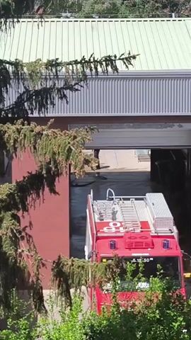 A red fire truck is seen from an elevated perspective, either backing into or pulling out of a large garage bay. The garage is part of a building with a red wall and a grey corrugated metal roof. A person is briefly visible in the background, walking inside the garage bay. The fire truck has equipment on its roof and a white number "5.18.50" printed on its front bumper. Lush green trees and shrubs are prominent in the foreground and around the building, partially obscuring the view. The scene appears to be set during daytime under bright, sunny weather. The overall impression is an active fire station or garage area, possibly in Vilafranca del Penedès, Spain.