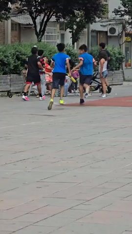 The video shows a group of children and adults in what appears to be a public outdoor space with a playground. In the foreground, a paved area with red markings is visible. In the background, there is a playground with a yellow slide, a swing set, and other play equipment. Several people are present in the video: two young boys in blue shirts are running, one of whom is holding a yellow object that resembles a bat. Other individuals, including adults and children, are gathered near a bench and a stroller. The environment seems to be urban, with buildings and trees visible in the background. The lighting suggests it is daytime, likely with overcast skies.