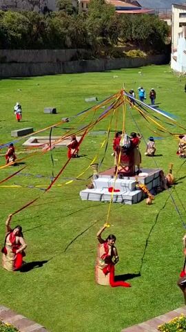 A high-angle, outdoor shot shows a traditional ceremony or performance on a bright green lawn. In the center, a group of people in colorful attire stands around a multi-tiered white and grey structure, pulling on ribbons of various colors that extend outwards. Several individuals in similar attire are positioned around the lawn, each holding a ribbon that stretches towards the central structure. The individuals in the foreground are kneeling on one knee, holding their ribbons high. In the background, other people are observing the event. The environment features a stone wall bordering the lawn and buildings in the distance.