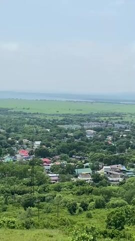 A high-angle shot, looking down on a rural town nestled in a valley surrounded by lush green trees and fields. The town consists of houses with red and green roofs, as well as some larger apartment buildings. In the distance, a road winds through the fields, leading to a range of hazy mountains under a bright, partly cloudy sky. The vegetation is dense and vibrant, indicating a temperate climate. There are no people or visible vehicles in the frame. The camera pans slightly to the right, revealing more of the expansive landscape.