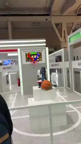 A person walks in front of a basketball game booth. The booth has a robot that is throwing a basketball. The robot successfully throws the basketball into a hoop. A woman is walking past the booth. The booth is in a large, open convention center.