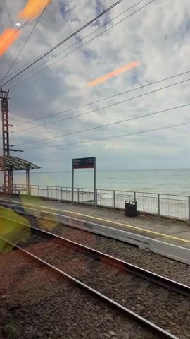 The video pans across a train station platform next to the ocean. On the platform, there are train tracks, a yellow safety line, a white fence, a trash can, and a bench. The ocean is visible beyond the fence, with a ship in the distance. The sky is cloudy. The video is shot from inside a moving train.