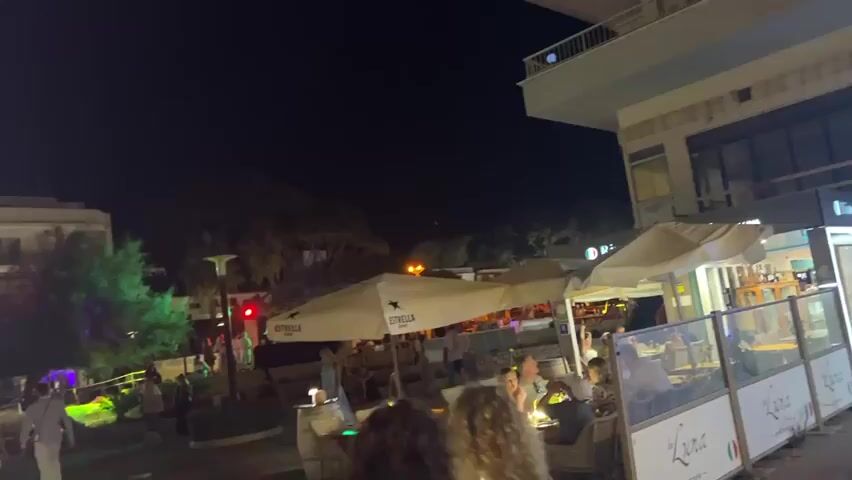 The video shows a nighttime view of a bustling street scene with people walking and dining outdoors. There are several buildings with illuminated storefronts and balconies. Some buildings are decorated with colorful lights, including green and purple hues. Large white umbrellas, some with logos, are set up outside establishments, suggesting outdoor seating areas for restaurants or cafes. People are seen walking along the sidewalk, and some are seated at tables, appearing to be engaged in conversation or enjoying their meals. The overall atmosphere seems lively and social. The camera pans and moves, giving a sense of walking through the area.